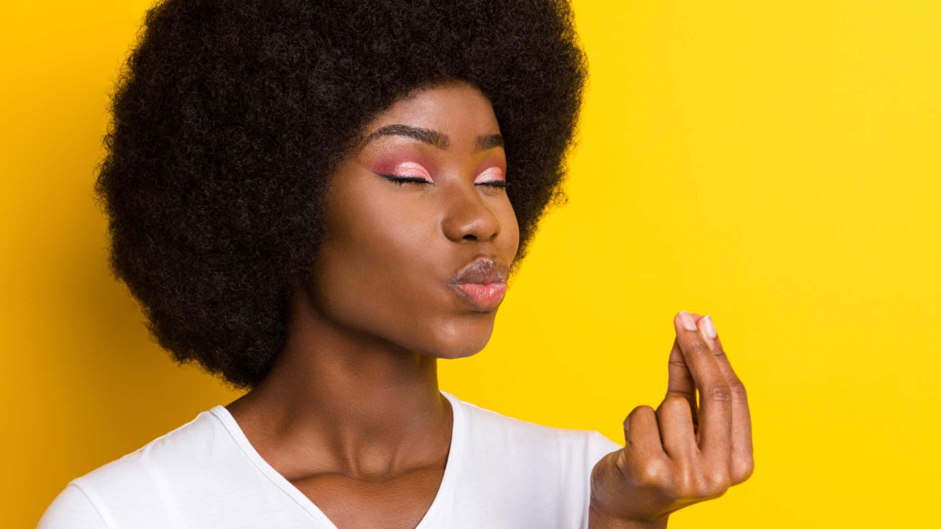 A dark-skinned woman with closed eyes and pursed lips holding her forefingers together against a yellow background