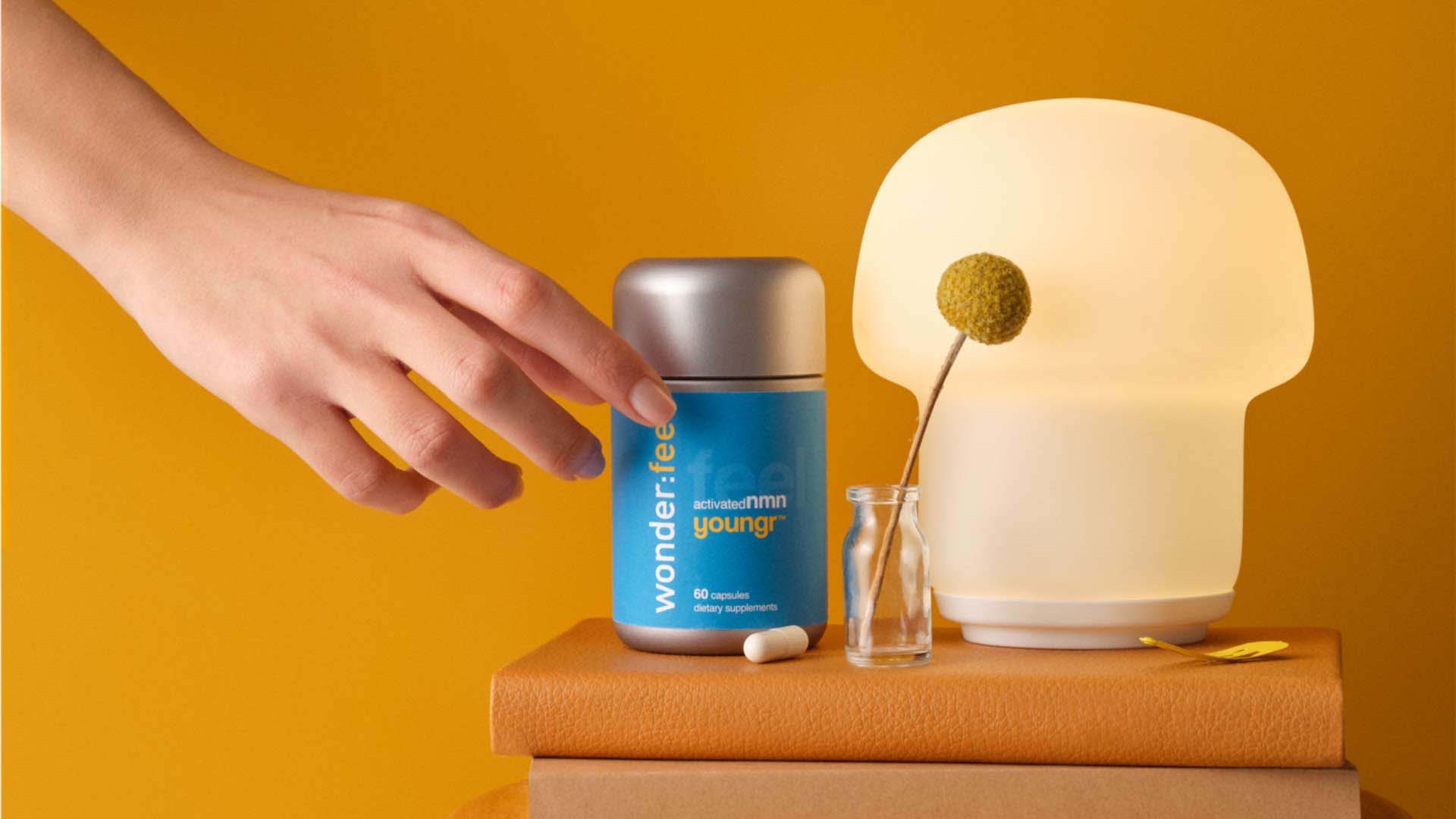 Hand reaching for a Youngr NMN bottle next to a small light, both set atop a stack of leather-bound books
