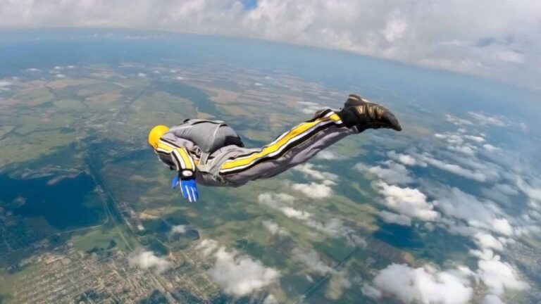 A skydiver in a yellow helmet and jumpsuit freefalls over a scenic landscape of fields and clouds, capturing the thrill of the dive and the spirit of wellness and longevity